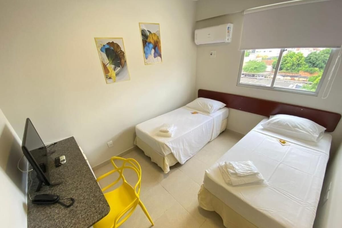 Ein kleines Hotelzimmer mit zwei Einzelbetten, einem Schreibtisch mit Computer, einem Stuhl und einem Fenster mit Blick auf Gebäude.