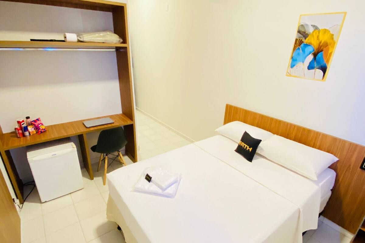 Ein Hotelzimmer mit Bett, Schreibtisch mit Stuhl, kleinem Kühlschrank und einem Regal mit verschiedenen Gegenständen.