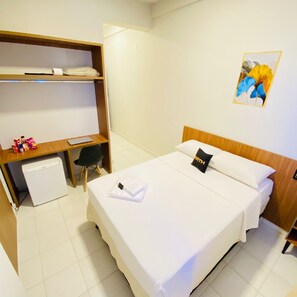 A hotel room with a bed, a desk with a chair, a small refrigerator, and a shelf with various items. - NORTH HOTEIS (Montes Claros)