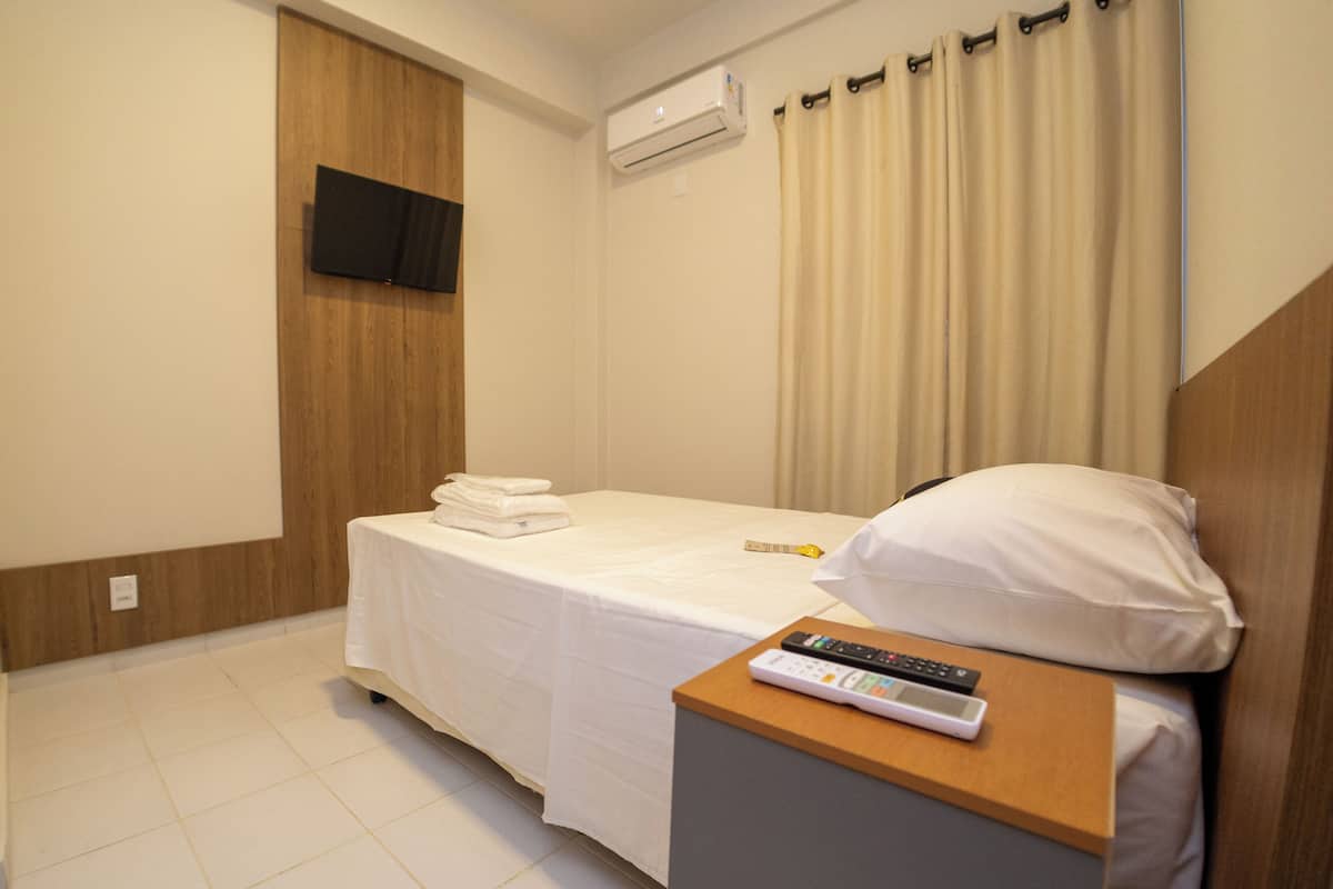 Ein Hotelzimmer mit einem Bett, einem Nachttisch mit Fernbedienung, einem an der Wand montierten Fernseher und einer Klimaanlage.