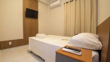 Quarto de hotel com uma cama, mesa de cabeceira com controle remoto, TV na parede e aparelho de ar condicionado.