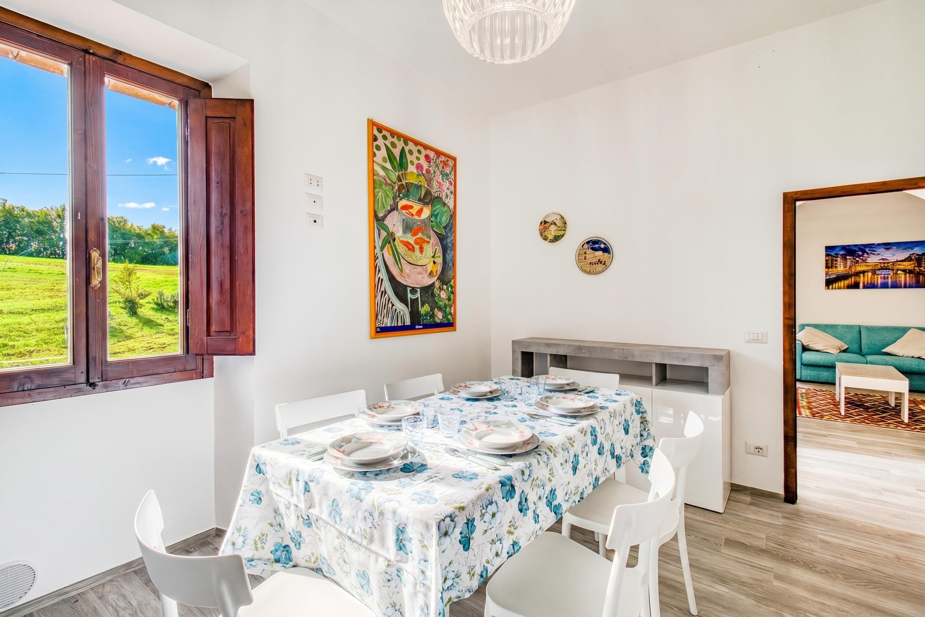 Ein Essbereich mit Tisch für vier Personen, weißen Stühlen, einer Tischdecke mit Blumenmuster und Blick auf ein grünes Feld durch ein Fenster.