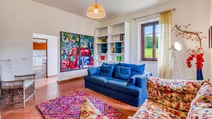 A living room with a blue sofa, a colorful rug, a bookshelf, and a large painting. - La Casa Della Quercia Relax In Tuscany (Montaione)