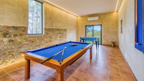 Une table de billard dans une pièce aux murs en pierre, avec une fenêtre et une vue sur la verdure à l’extérieur.