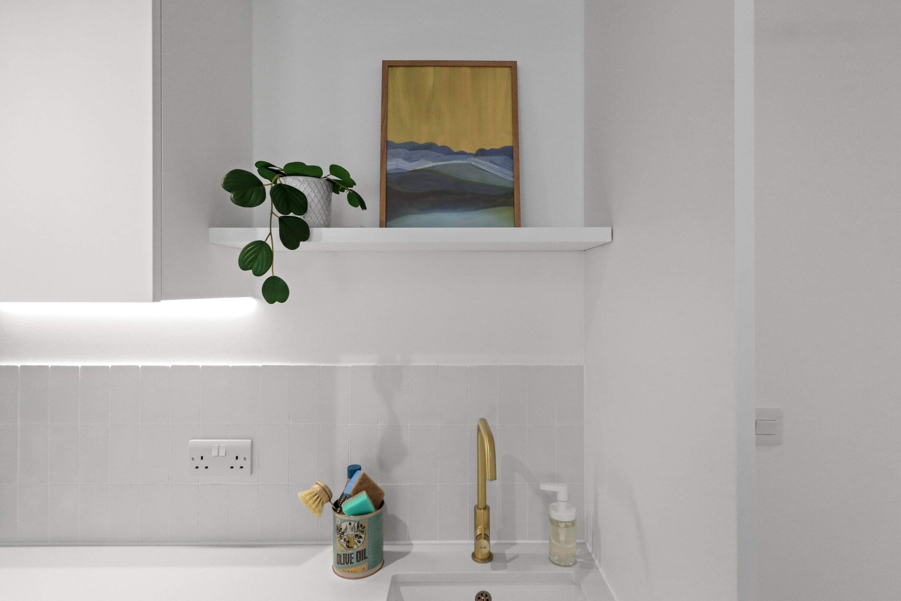 Casa de banho com lavatório branco, torneira dourada, planta, obra de arte emoldurada e um recipiente com pincéis e dispensador de sabonete.