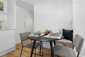 A modern dining area with a round table, grey upholstered chairs, and a small side table with a bowl of fruit. - Relax in Style Elegant Apartment in Eastbourne (Eastbourne)