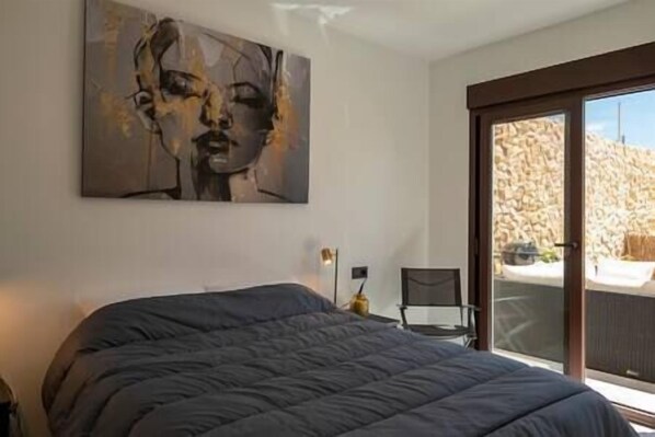 A bedroom with a large bed, a chair, a painting, and a view of a stone wall through a sliding door.