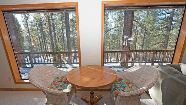 Une pièce avec une table ronde en bois et deux chaises en osier agrémentées de coussins colorés, de grandes fenêtres donnant sur une forêt et une terrasse.