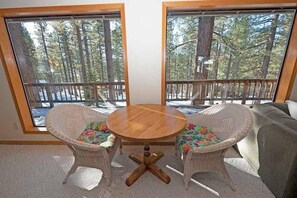 A room with a round wooden table and two wicker chairs with colorful cushions, large windows with a view of a forest and a deck. - 2293 Marshall Trail 4 Bedroom Home by RedAwning (South Lake Tahoe)