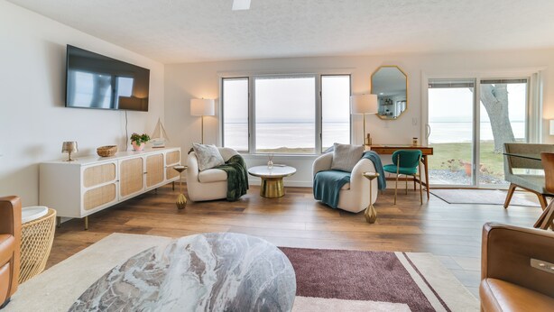 A spacious living room with a large window offering a view of the sea, a ceiling fan, and a flat-screen TV mounted on the wall. - Bright Houghton Lake Home w/ Boat Dock + Fire Pit (Houghton Lake)