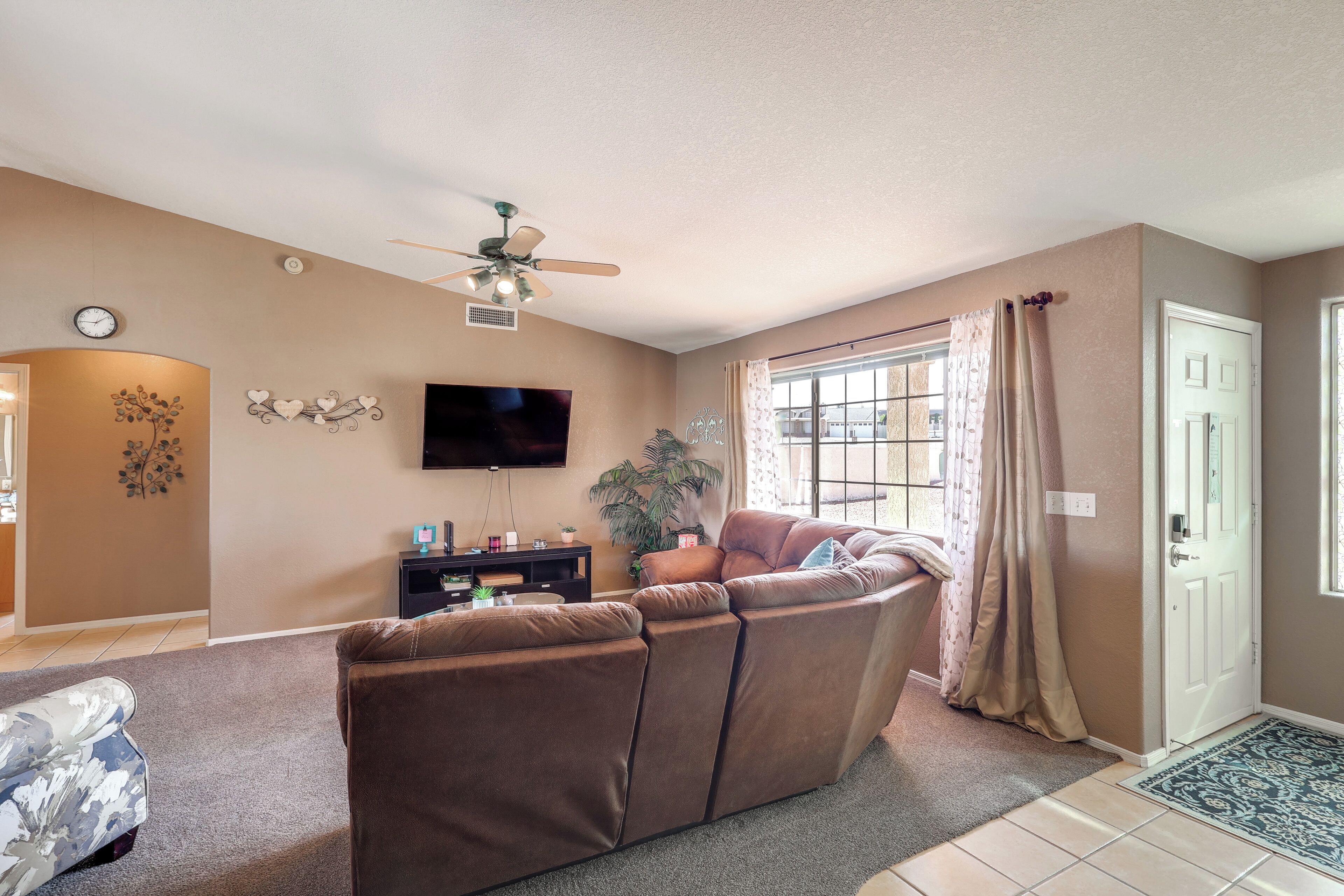 A living room with a brown sectional sofa, a flat-screen TV mounted on the wall, a ceiling fan, and a window with curtains.