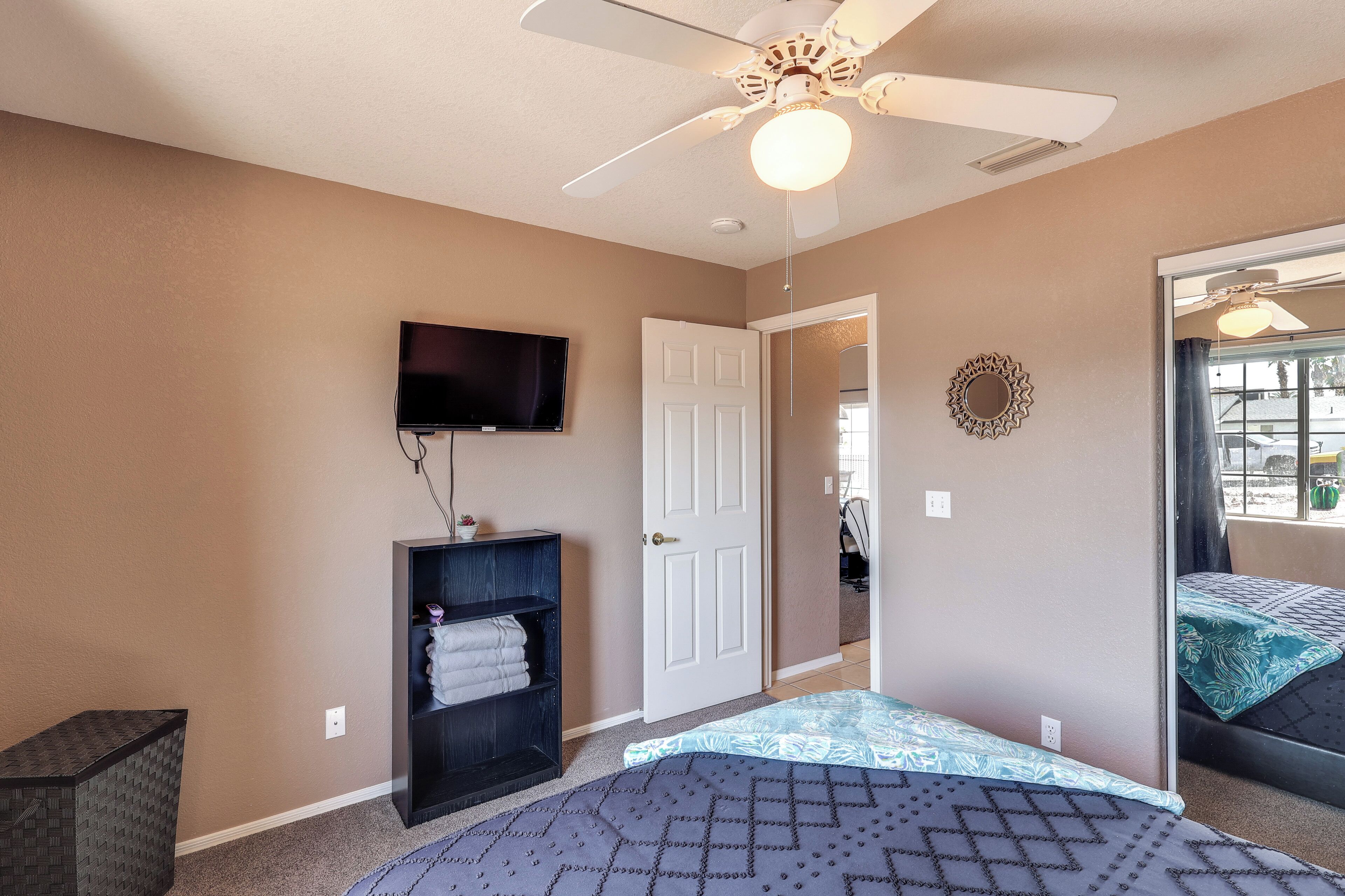 A bedroom with a bed, a TV mounted on the wall, a ceiling fan, and a door leading to another room.
