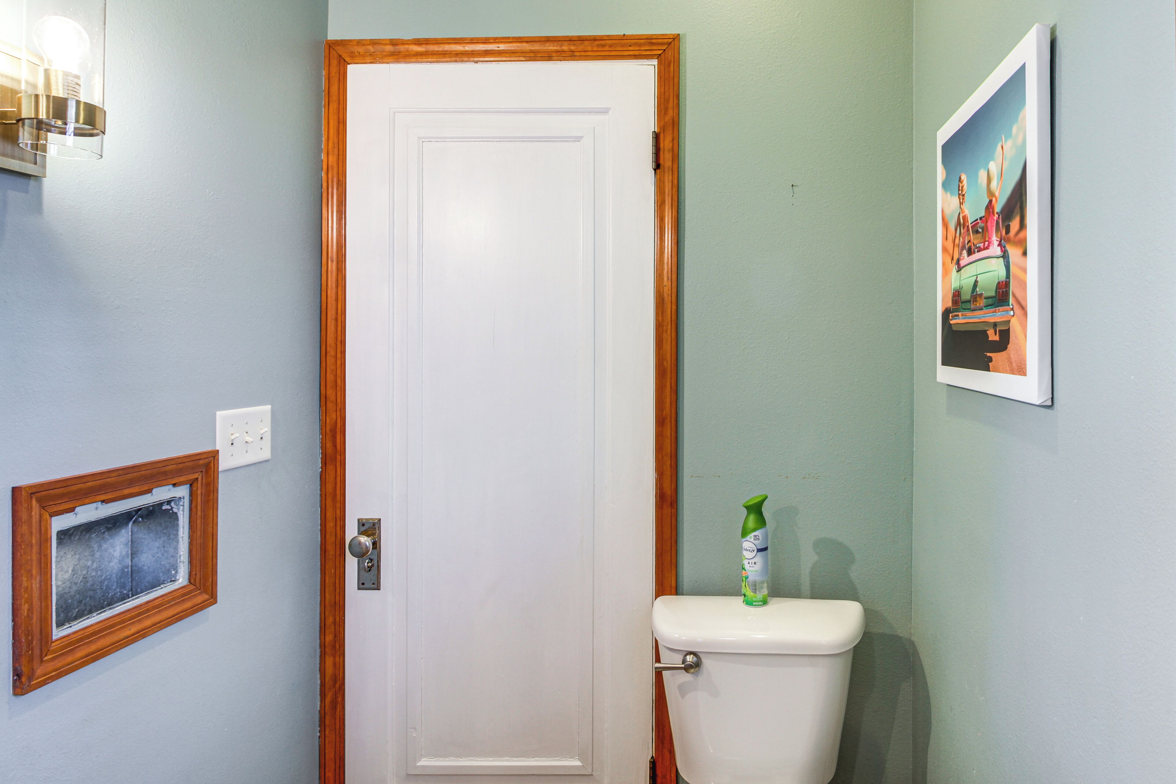 A bathroom with a white door, a toilet, a framed window, and a wall-mounted light fixture.