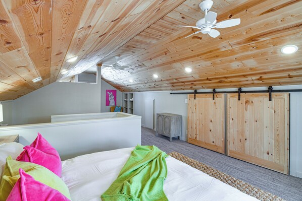 A bedroom with a wooden ceiling, a bed with colorful pillows, a ceiling fan, and a small cabinet.