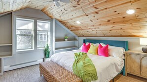 A bedroom with a sloped ceiling, a bed with colorful pillows, a bedside table, and a window with a view of trees. - Spacious Detroit Lakes Home: Walk to Downtown! (Detroit Lakes)