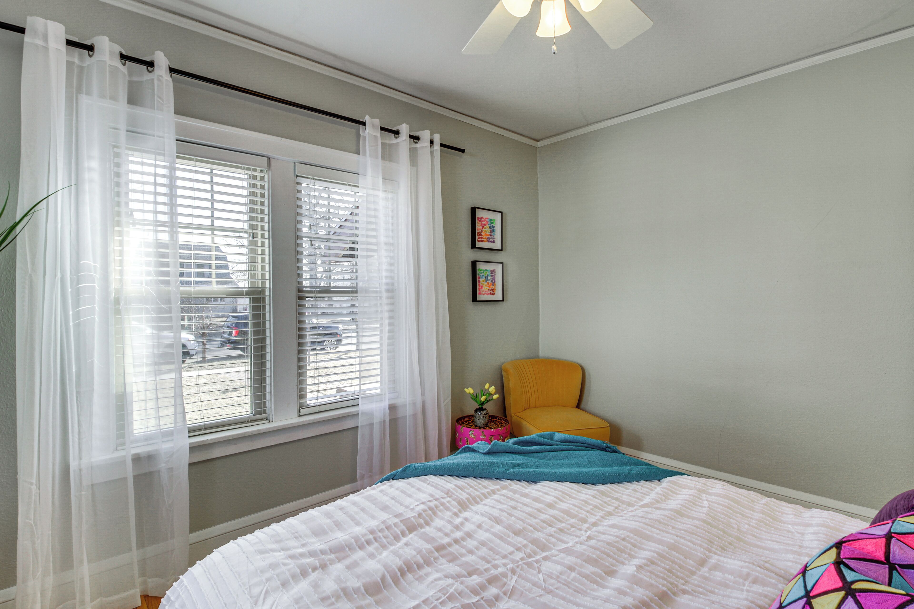 A bedroom with a bed, a window with sheer curtains, a chair, and two framed pictures on the wall.