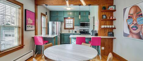 Una cocina moderna con armarios azul verdoso, encimera blanca y una isla central. Hay un comedor con una mesa redonda y sillas.