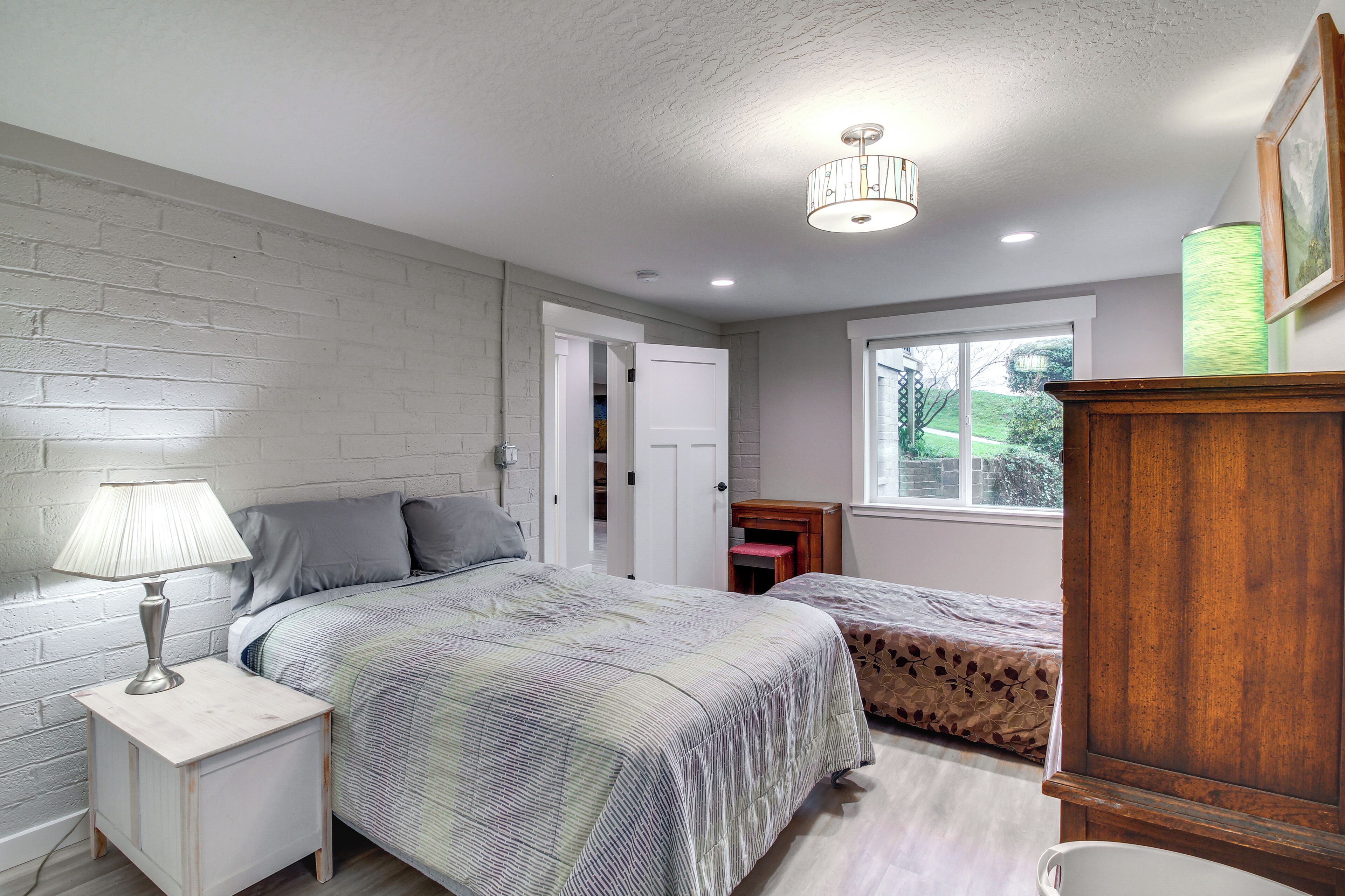 A bedroom with two beds, a nightstand with a lamp, a wooden cabinet, and a window with a view of greenery.