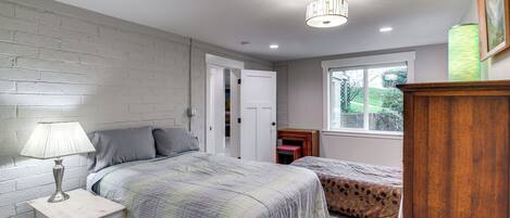 A bedroom with two beds, a nightstand with a lamp, a wooden cabinet, and a window with a view of greenery.