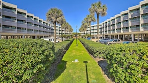 A hotel corridor with a central pathway flanked by neatly trimmed hedges and palm trees, leading to a building with multiple balconies. - Galveston Condo w/ Beach Access Near Moody Gardens (Galveston)