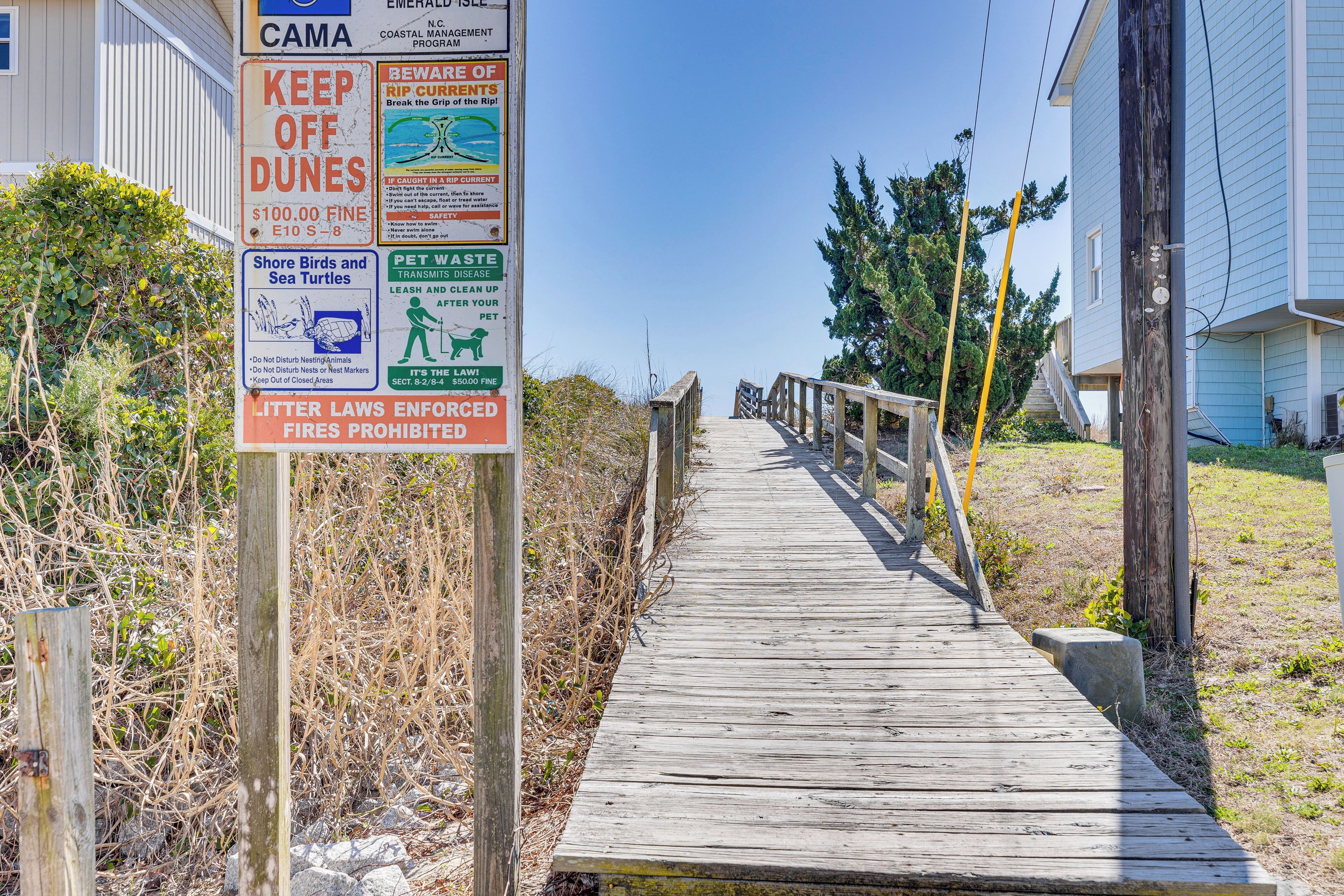 Un muelle de madera que conduce a una casa con varios carteles, incluyendo uno sobre no pisar las dunas y una multa por tirar basura.