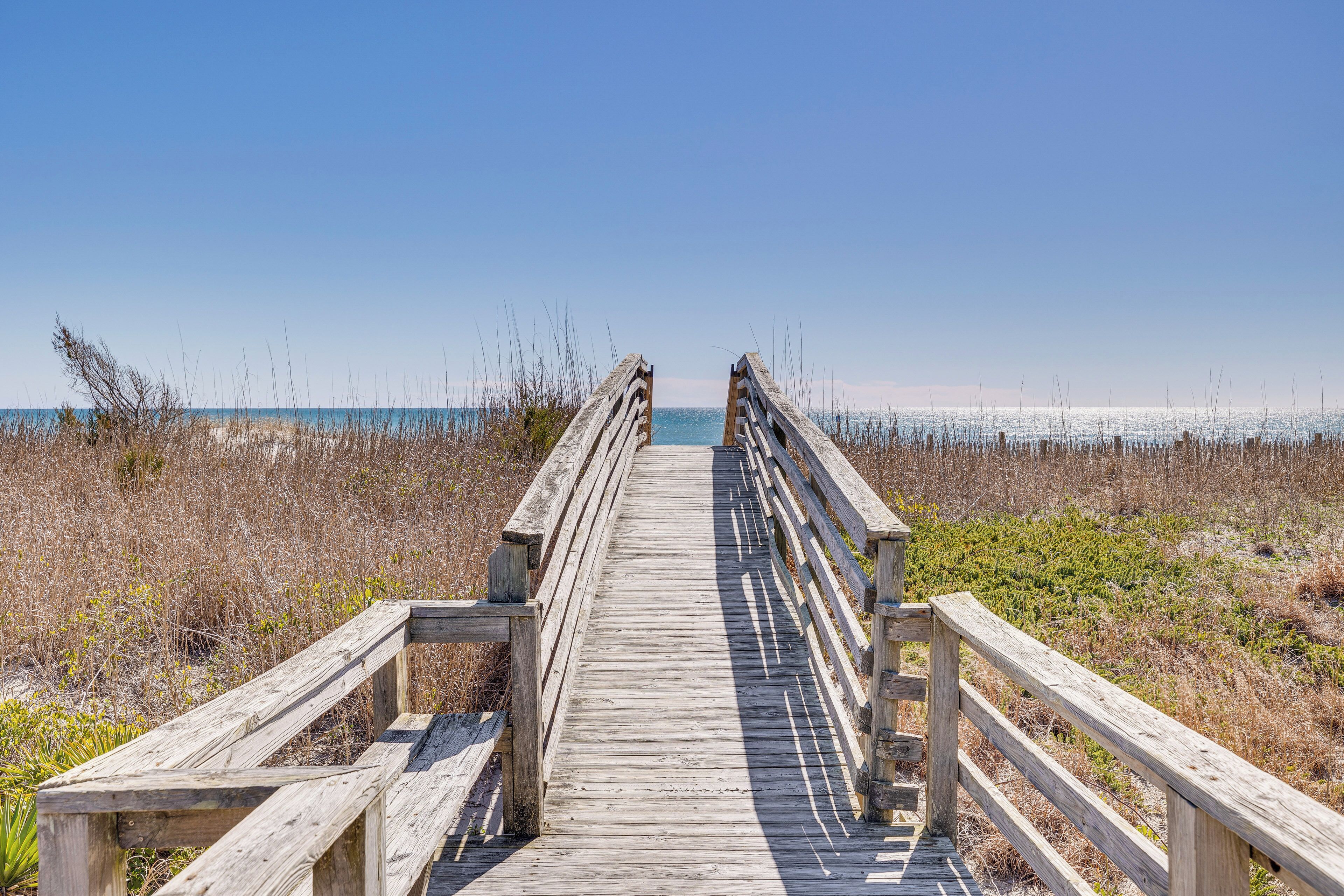 A wooden boardwalk leading to the beach, surrounded by dry grass and shrubs.