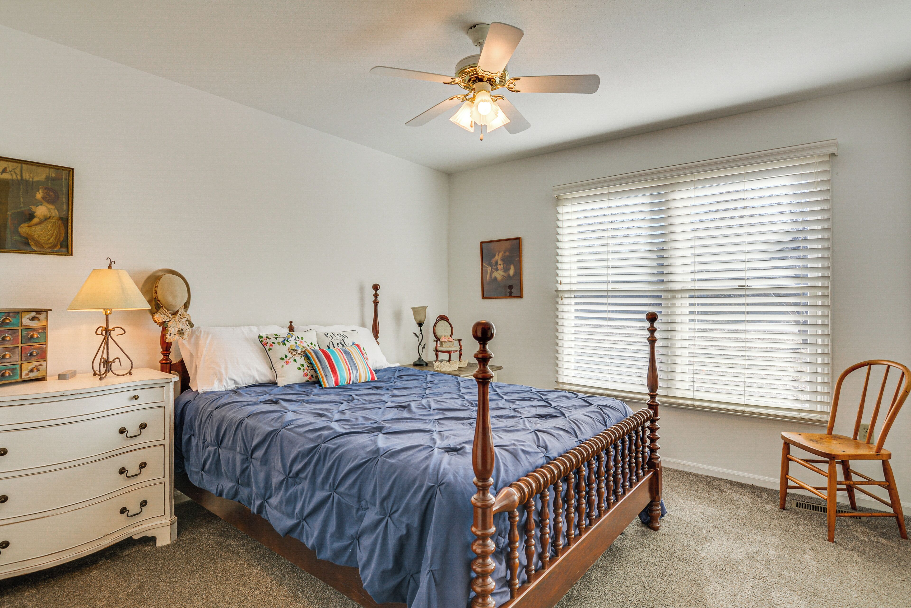 A bedroom with a bed, dresser, chair, ceiling fan, and window with blinds.