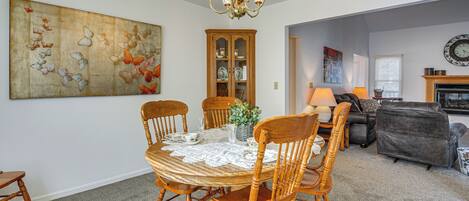 Un coin repas avec une table ronde en bois, quatre chaises, un lustre, un grand tableau représentant un papillon, un meuble avec des portes en verre et une cheminée en arrière-plan.