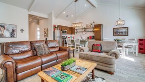 A spacious living room with a brown leather sofa, a beige armchair, and a wooden coffee table. There is a kitchen area with wooden cabinets and a dining table in the background. - Idyllic Lago Vista Home < 1 Mi to Lake Travis! (Leander)