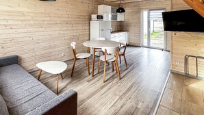 A modern, compact living space with a wooden interior, a dining area with white round tables and chairs, a kitchenette, and a television mounted on the wall.