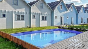 A swimming pool with a wooden deck, surrounded by a grassy area and a row of houses with gabled roofs.