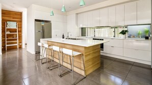 A modern kitchen with white cabinets, a central island with bar stools, and pendant lighting. - Mount Coolum Beachfront Abode with Private Rooftop (Coolum Region)