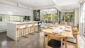 A modern kitchen with a central island, bar stools, and a dining area with a long table set for six. - Mount Coolum Beachfront Abode with Private Rooftop (Coolum Region)