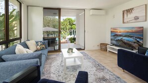 A living room with a sofa, a coffee table, and a TV. There is a sliding glass door leading to an outdoor area with plants and a patio.