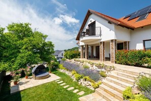 A two-story house with a balcony, a stone pathway, and a garden with various plants. - Pilger Apartments (Tihany)