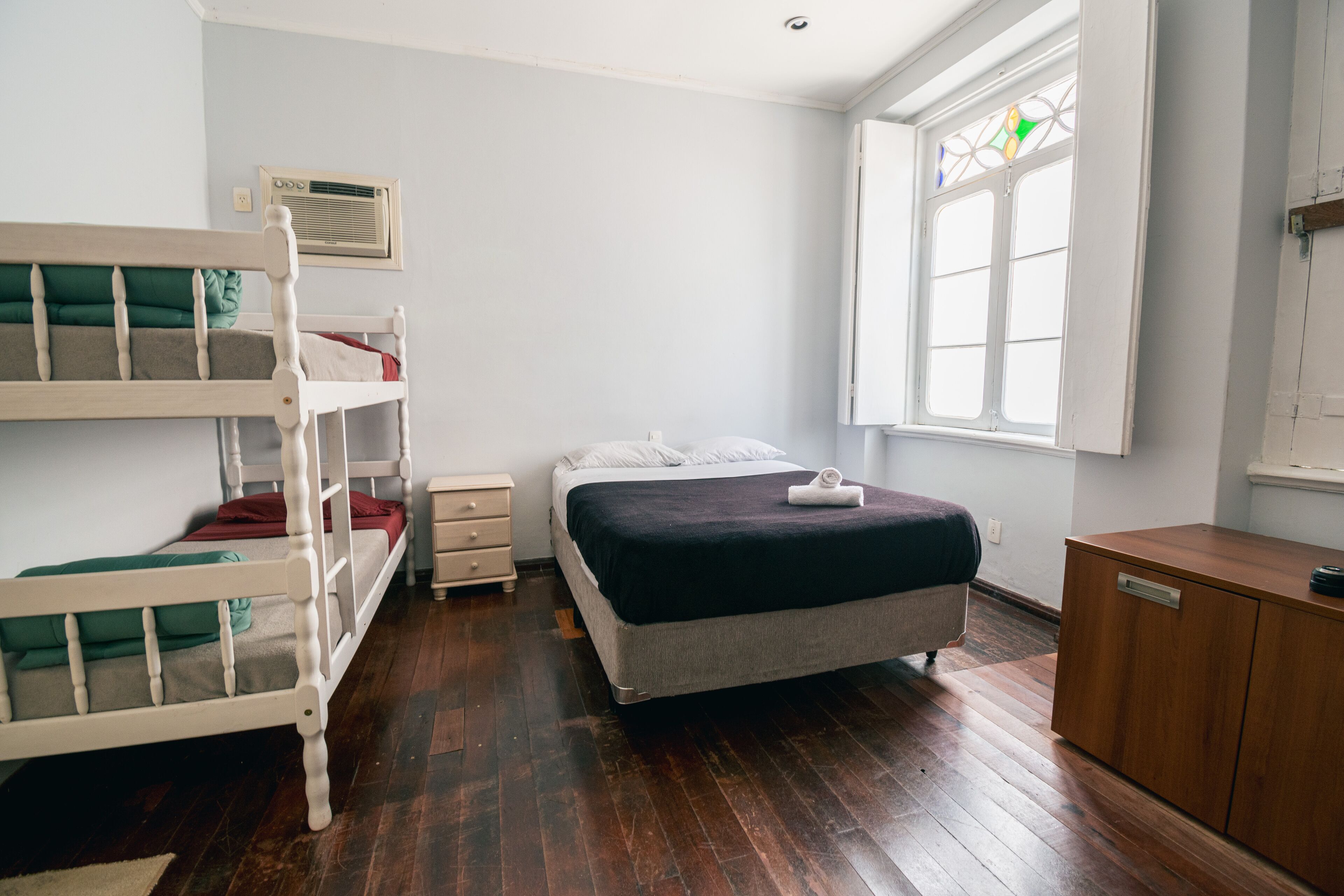 A room with bunk beds, a single bed, a wooden cabinet, and a window with a stained glass design.