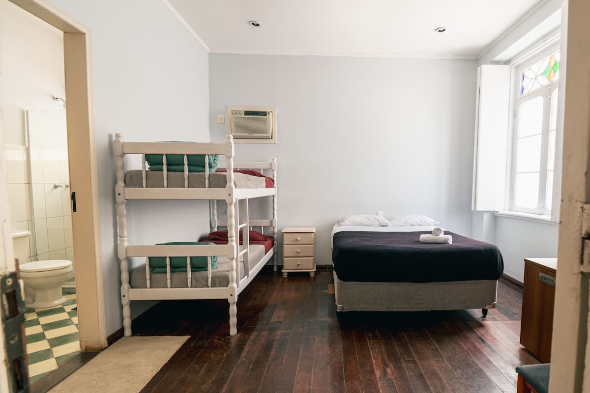 A room with a bunk bed, a single bed, a bedside table, a nightstand, a window, and a door leading to a bathroom.