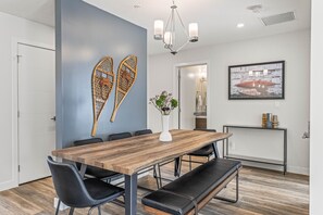 Un comedor moderno con una mesa de madera, sillas negras y un banco. Hay un florero con flores sobre la mesa, y una lámpara de pared encima.