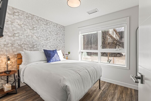 A bedroom with a large bed, a window with a view of the outside, a bedside table with a lamp, and a wall with a floral pattern.