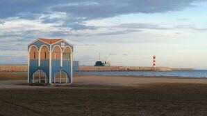 Une plage avec une petite construction bleue et blanche, un phare et une digue rocheuse.