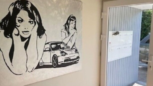 Ein Zimmer mit einer zur Balkontür führenden Tür, einem an der Wand befestigten Kunstwerk mit einem Auto und einer Frau sowie einem mit Jalousien versehenen Fenster.