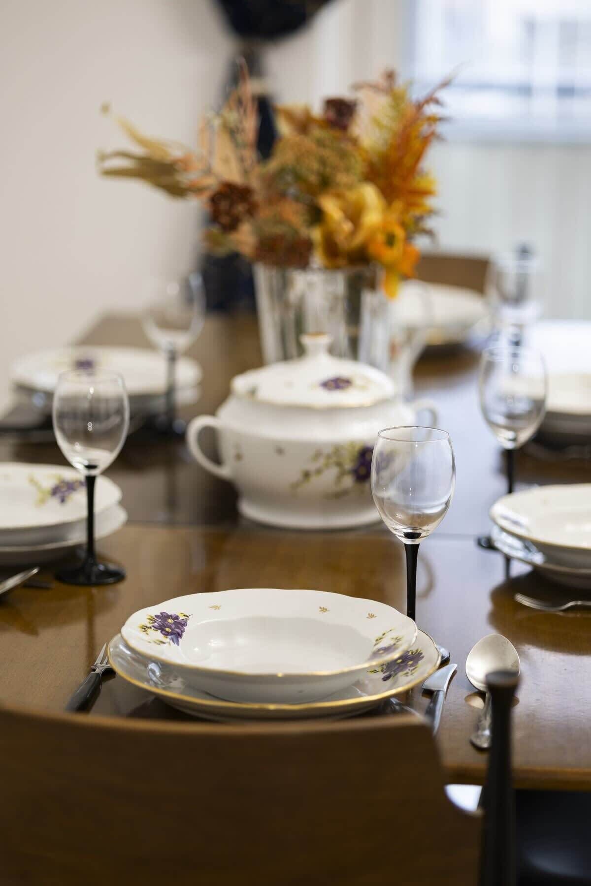 Een gedekte eettafel met borden, glazen en bestek, met een bloevas op de achtergrond.