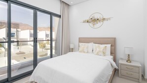 Quarto moderno com uma janela grande, cama com roupa de cama branca, cabeceira, mesas de cabeceira e um objeto decorativo na parede.
