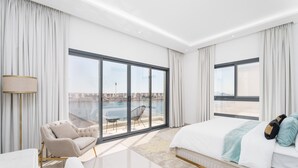 Quarto de hotel moderno com uma cama grande, área de estar com uma poltrona e vista para um corpo de água através de janelas do chão ao teto.