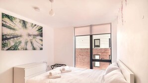 A modern bedroom with a large bed, a desk, and a view of a brick building through a window. - Stunning Modern 1 Bed Apartment, Manchester (Salford)