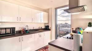A modern kitchen with white cabinets, a black microwave, a coffee maker, and a view of the city through a large window. - Stunning Modern 1 Bed Apartment, Manchester (Salford)