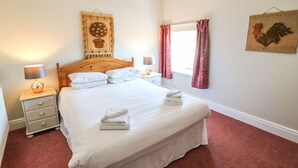 Un lit bien fait, avec du linge de lit blanc et une serviette pliée posée sur le côté. On y trouve une tête de lit en bois, une table de chevet avec une lampe, et une œuvre d’art accrochée au mur au-dessus du lit.