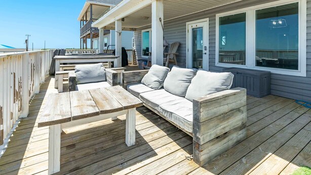A wooden deck with a sofa and a table, overlooking a building with large windows. - SeaScape Haven - Right on the sandy BEACH, Hot Tub! (Surfside Beach)