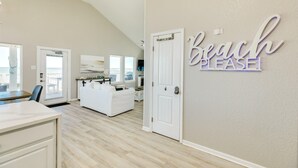 A spacious living room with a high ceiling, a white couch, a dining area with a table and chairs, and a door with a sign reading "Beach Please!". - SeaScape Haven - Right on the sandy BEACH, Hot Tub! (Surfside Beach)