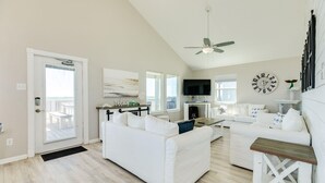 A spacious living room with a white sofa, a coffee table, and a TV. There is a door leading to a balcony, and a ceiling fan is visible. - SeaScape Haven - Right on the sandy BEACH, Hot Tub! (Surfside Beach)
